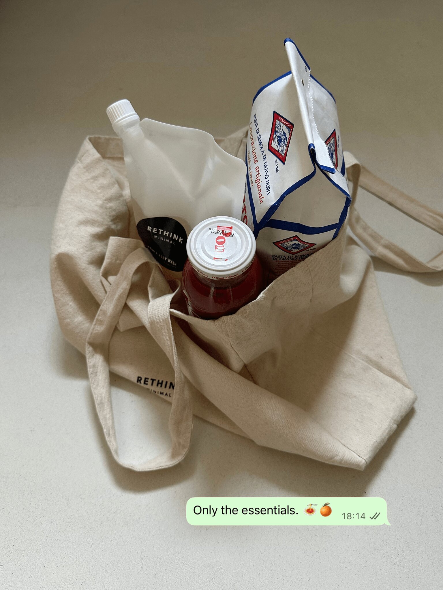 Stofftasche auf beigem Mikrozement mit Rethink Minimal Hand & Body Wash Refill, Pasta & Tomatensauce. Darunter ist eine WhatsApp Nachricht mit den Worten "Only the essentials."